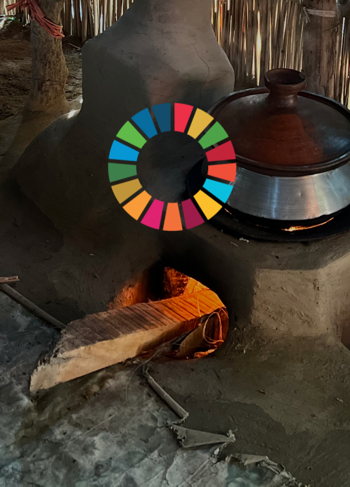 Clay oven with a metal pot on top, glowing fire inside opening, wooden plank bridge to oven, rustic outdoor setting with straw walls and tools nearby.