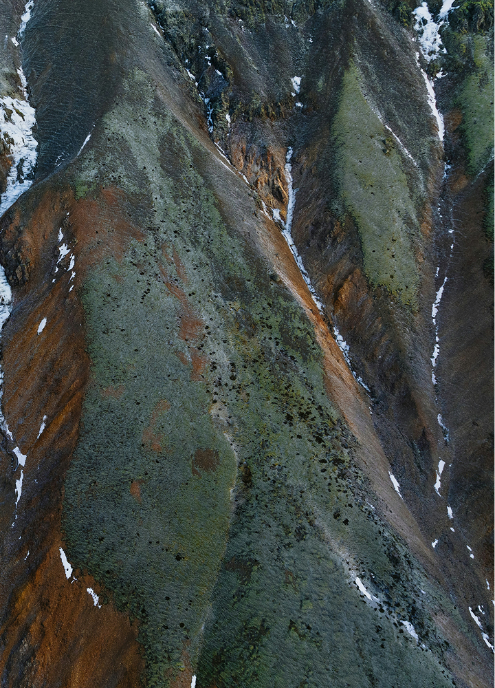 Aerial view of a rugged mountainside with multicolored rock faces in greens, browns, and grays, narrow snowy streaks, and sparse vegetation patches.