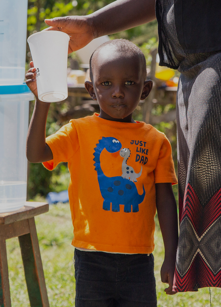 A young child in an orange shirt with dinosaurs holds a white cup up while an adult nearby offers a drink, outdoors with greenery in the background.