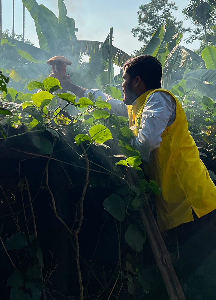 A person wearing a yellow jacket leans over a dark fence, reaching upward to touch a foggy, smoky area among lush green vines and banana leaves.