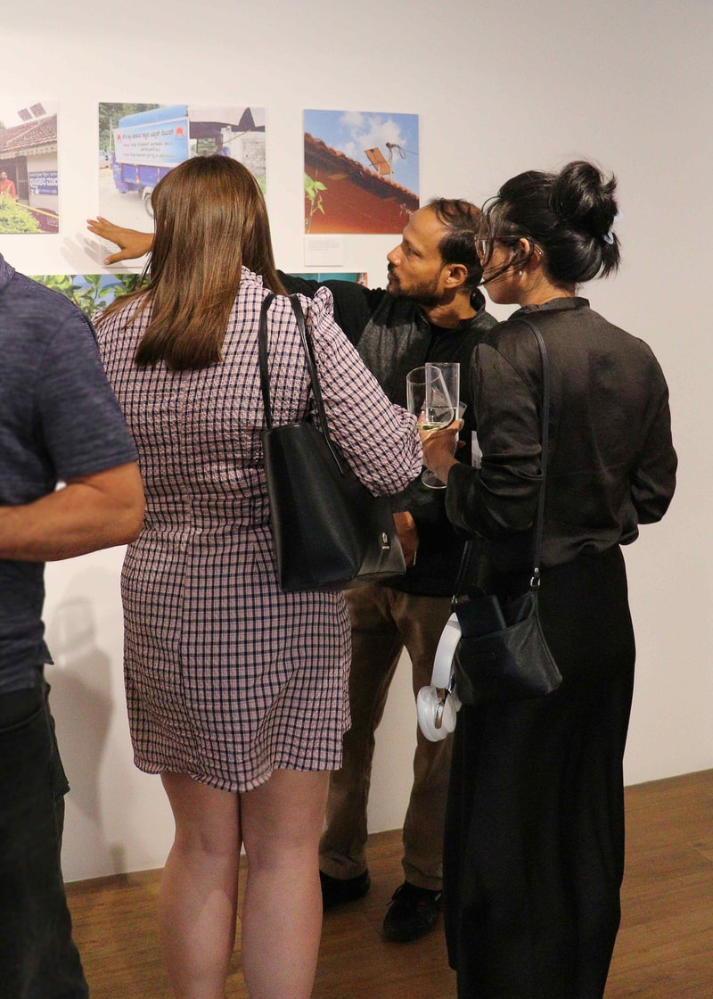 Three people stand at an art exhibit, looking at photos on the wall; one wearing a checked dress points, another holds a glass, and a third with a black outfit listens nearby.