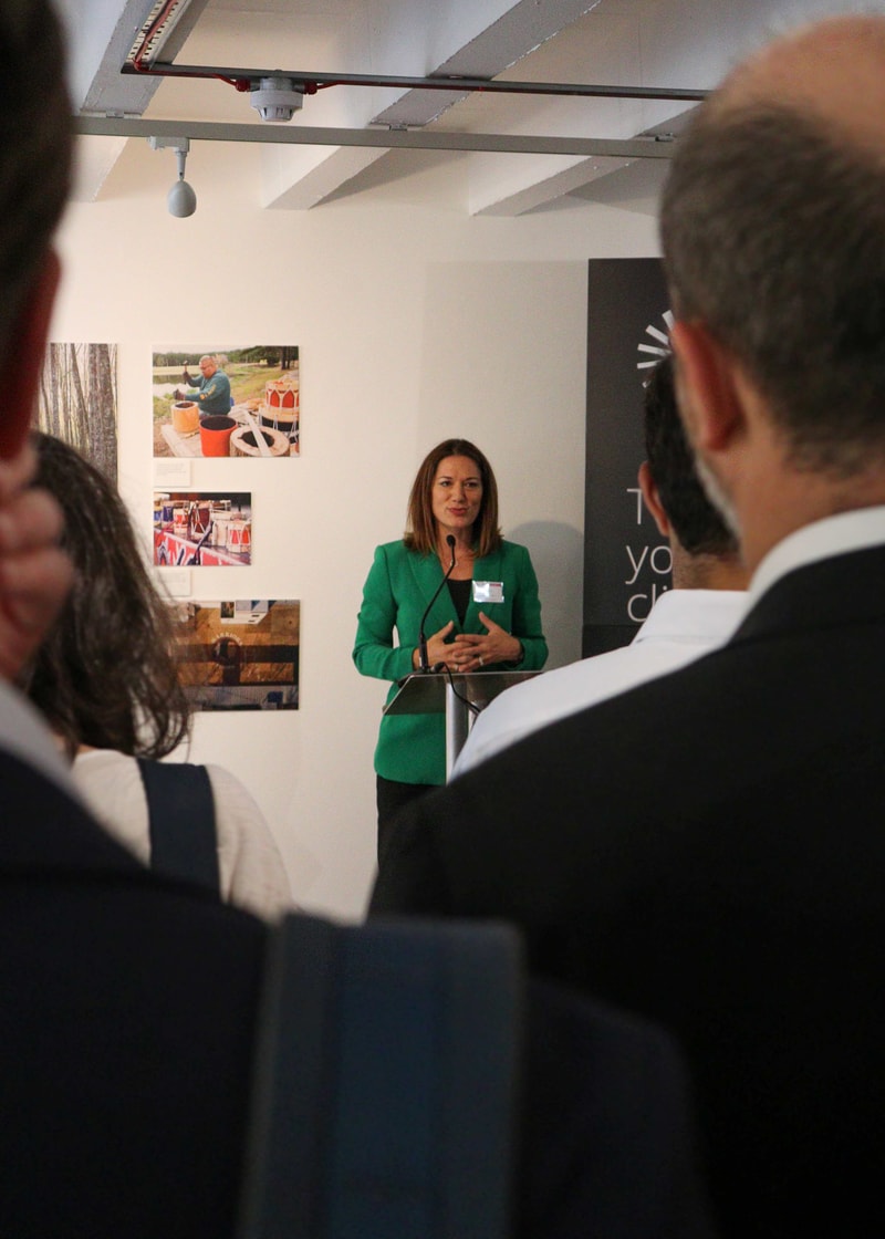 Speaker at podium in a green blazer addressing an audience in a gallery or conference space, with framed photos on the wall behind.