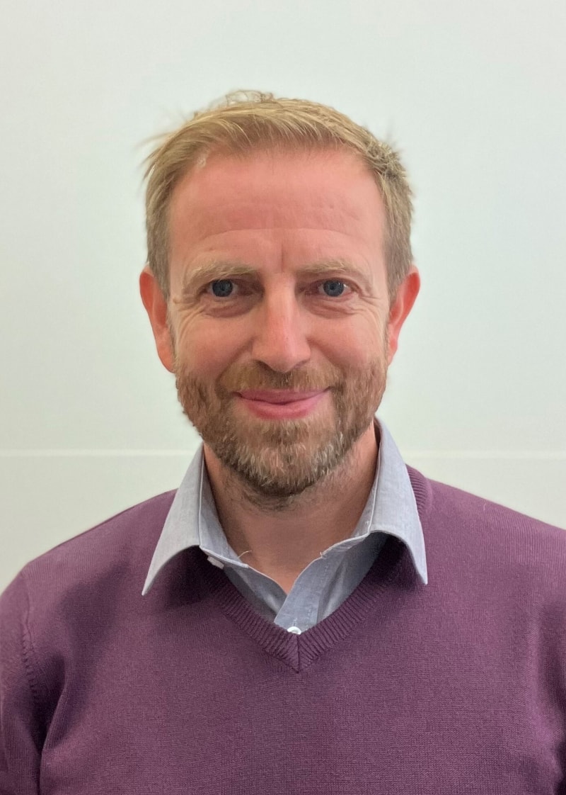 Portrait of a smiling man with short blond hair and a trimmed beard, wearing a light blue collared shirt under a maroon V-neck sweater, against a plain light background.