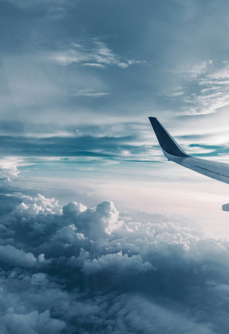 Wing of an airplane slicing through a blue sky above fluffy clouds, with a gentle horizon glow and distant cloudscape below.