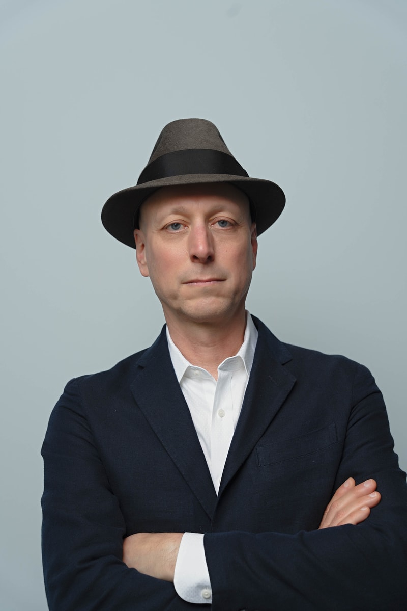 A person wearing a dark blazer and white shirt, with arms crossed, facing the camera, and wearing a brown fedora hat against a plain light gray background.