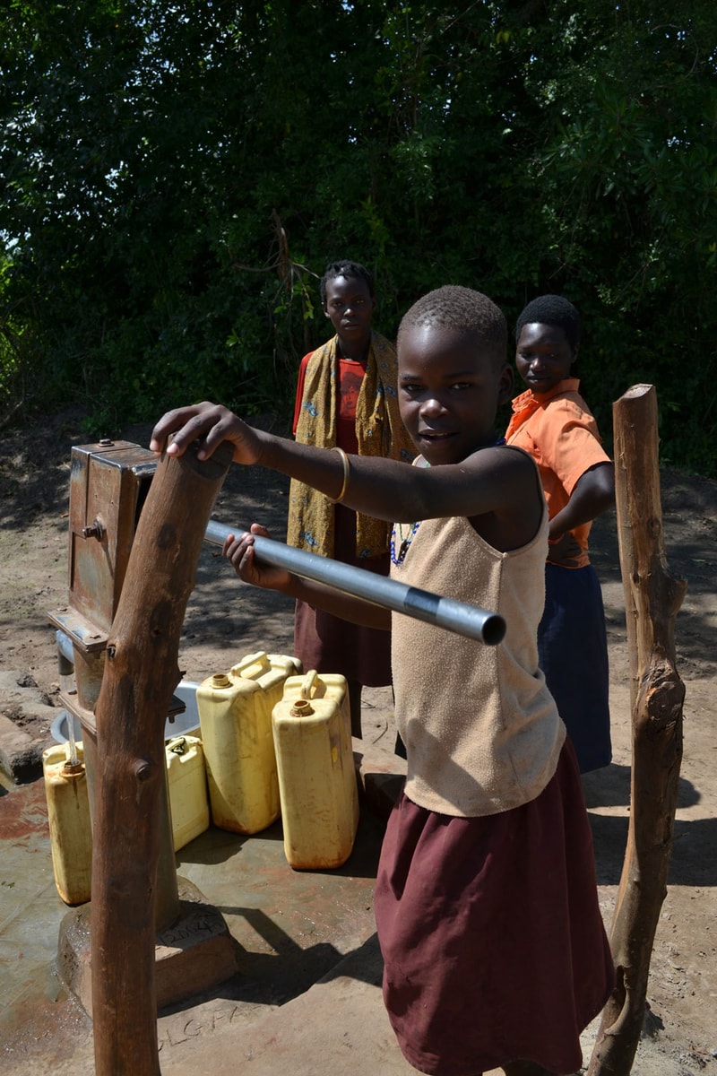Three children at a rural well, with a girl in a beige top and maroon skirt turning a metal pipe, while two others observe; yellow jerrycans and wooden posts nearby.