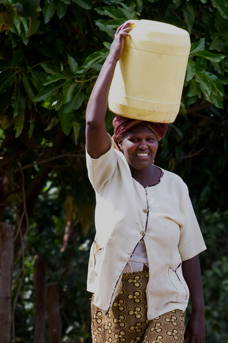 Woman balancing a large yellow water container on her head, smiling, wearing a light short-sleeve top and patterned skirt, outdoors with green foliage in the background.