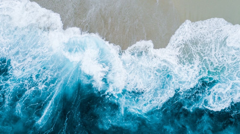 Aerial view of turquoise ocean waves crashing onto a sandy shore, white foam roiling where the sea meets the beach.