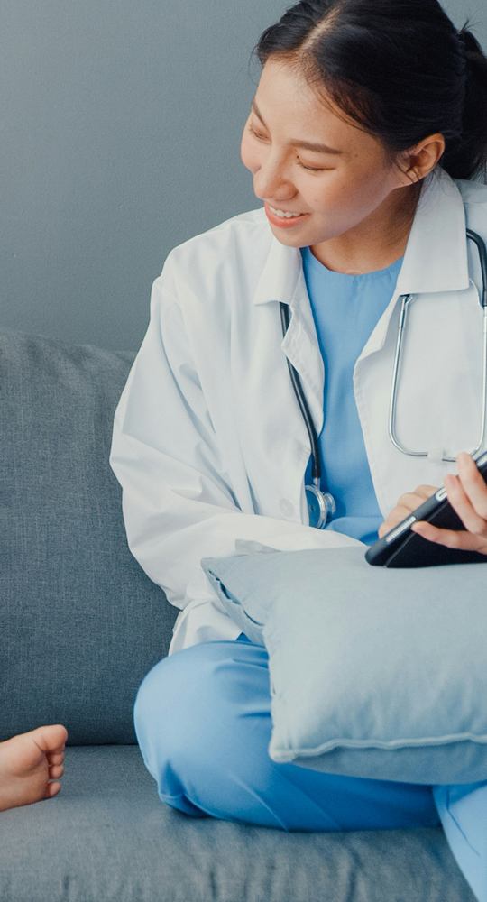 A healthcare professional in light blue scrubs and white coat sits on a sofa, smiling while holding a phone and a stethoscope rests around their neck.