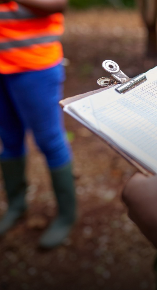 Clip-on clipboard with rings and paper held in foreground, blurred person in high-visibility orange vest and blue pants standing outdoors in a wooded area.