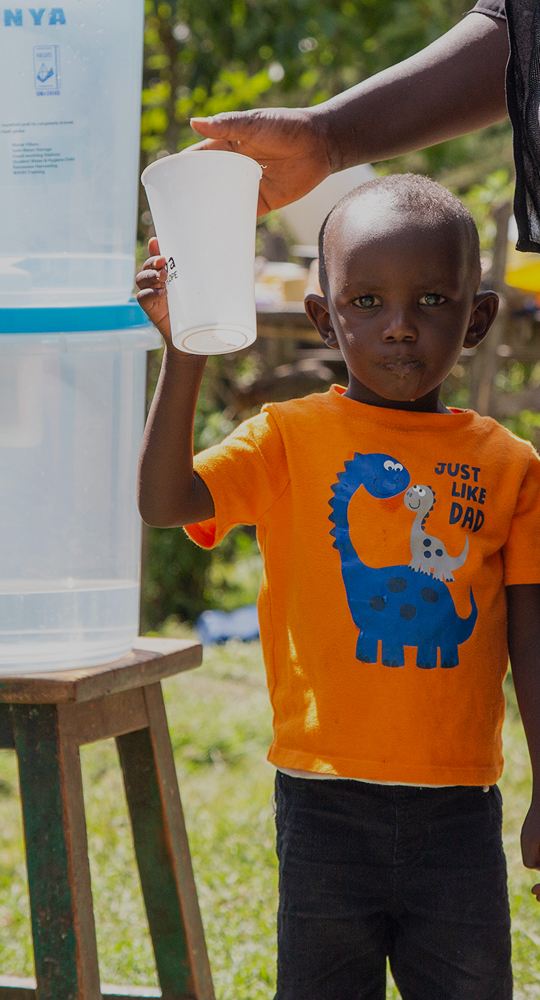 A young child wearing an orange shirt with a blue dinosaur print stands outdoors as an adult hands them a white cup near a large water cooler.
