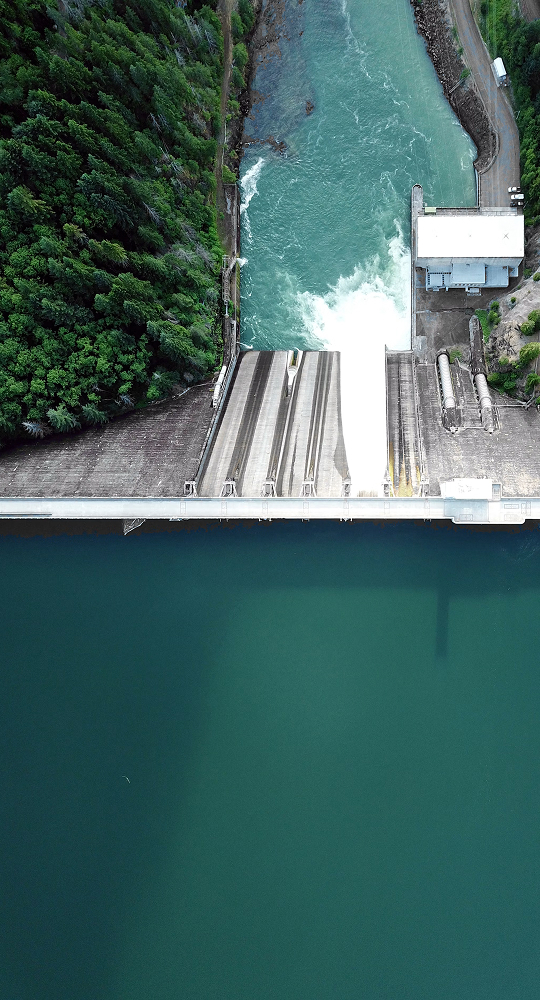 Aerial view of a dam with water pouring over spillways into a turquoise reservoir, surrounded by dense green trees and a roaded concrete structure at the top.