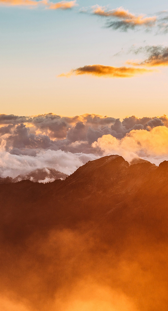 Sunlit mountain ridge rising above a sea of golden clouds at sunset, with warm orange haze and soft pink sky tones in the distance.