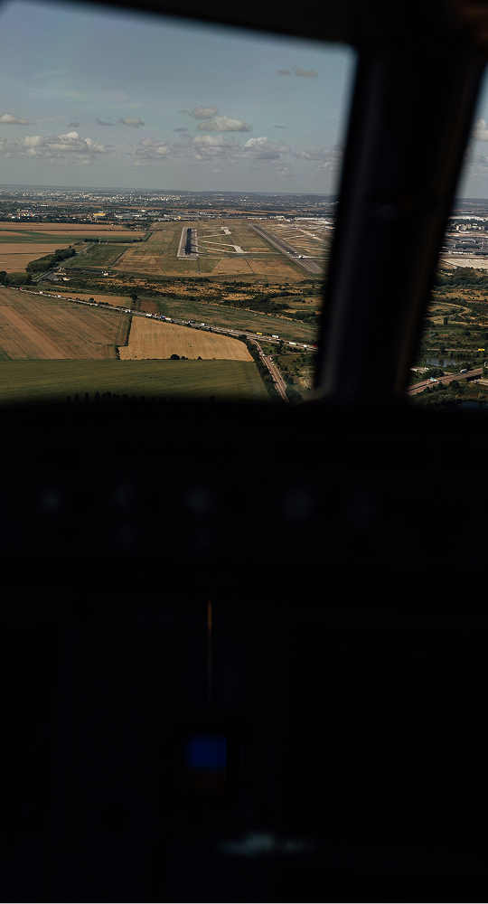 Aerial view of a countryside landscape with fields, roads, and a runway visible in the distance, framed by a dark airplane cockpit window.