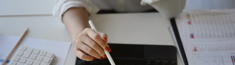 A close-up of a hand with orange nail polish gripping a white stylus over a keyboard, with papers and a chart in the background.