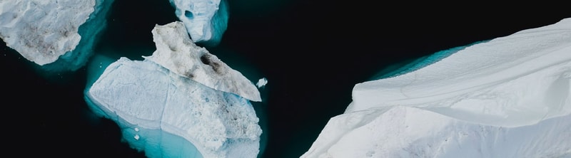 Aerial view of floating icebergs and a dark, inky body of water, with large white and blue-tinted ice chunks drifting near a snow-covered shore.