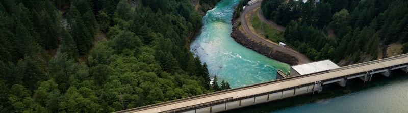 Aerial view of a turquoise river winding through a dense evergreen forest, with a paved road and concrete dam/bridge running along the water’s edge.