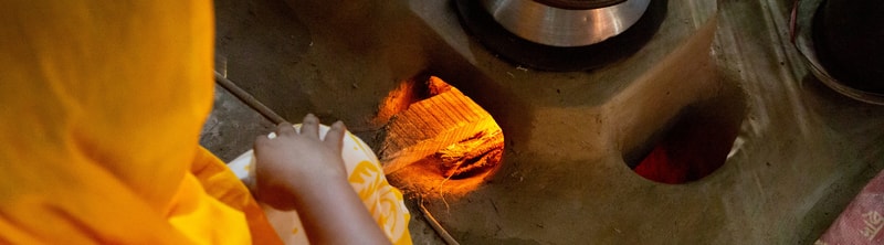 A person in a yellow garment feeds glowing orange-hot wood or embers into a furnace opening, with metal and charred surfaces around and a bright flame inside.