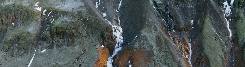 Aerial view of rugged, multicolored rocky mountains with patches of snow and green moss, showing deep crevices and steep slopes.