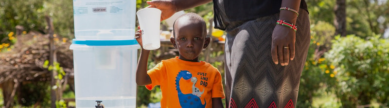 A child in an orange shirt with a dinosaur graphic stands beside an adult, who is holding a white cup over a large water dispenser outdoors.