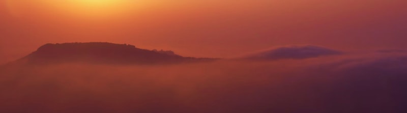 A hazy orange sunset over distant hills, with soft purple mist rolling across the lower landscape and a warm golden glow in the sky.