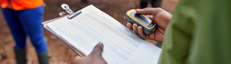 A person holds a clipboard with a printed spreadsheet and uses a handheld device, while another person in a green sleeve notes in the foreground outdoors.