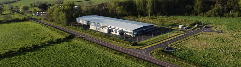A large modern building with a white roof sits along a curved road in a green rural area, surrounded by fields, trees, and a parking lot.