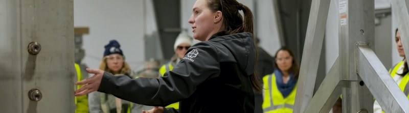A woman in a dark jacket gestures with her arms while a group of people in high-visibility vests watches inside an industrial facility with large metal structures.