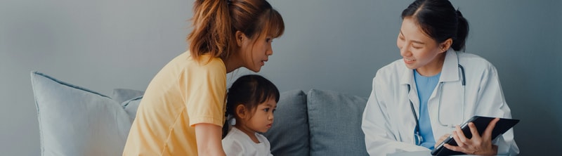 A healthcare professional in a white coat with a stethoscope sits with a young girl and a woman on a couch, discussing something on a tablet or clipboard.