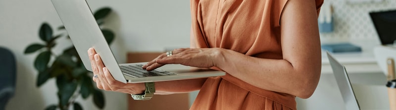 Person standing at a laptop, wearing a rust-colored sleeveless top and a light bracelet, typing with both hands in a bright office space.