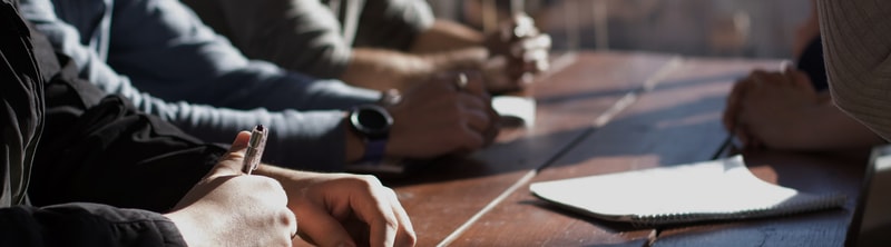 Several people sit at a long wooden table, writing with pens; close-up focus on a hand holding a pen in the foreground, with notebooks and blurred hands in the background.
