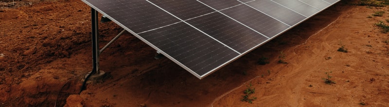 Solar panels mounted on metal supports tilted over red soil, with small green weeds and dusty ground extending into the distance.