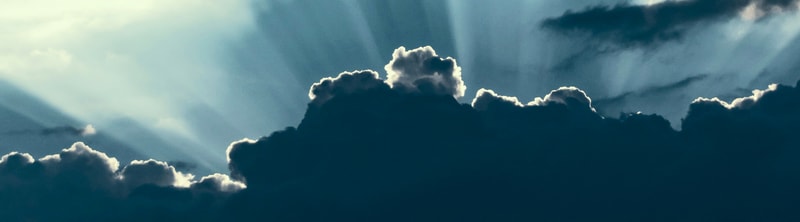 Bright sky with dark, heavy clouds and sun rays streaming through gaps, creating a dramatic contrast of light and shadow across the clouds.