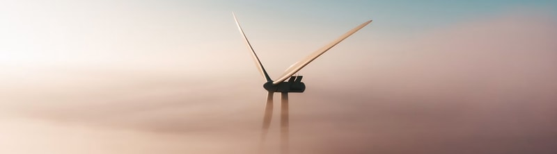 Wind turbine towering above soft, pastel clouds at sunrise, with long blades and a calm, misty horizon.