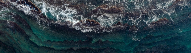 Aerial view of dark rocky coastline with turquoise ocean water below, white sea spray along jagged rocks, and vivid teal to deep blue gradients in the water.