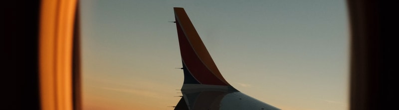 Silhouette of an aircraft tail viewed through a window, with warm orange light on left and soft gradient sky in the background.