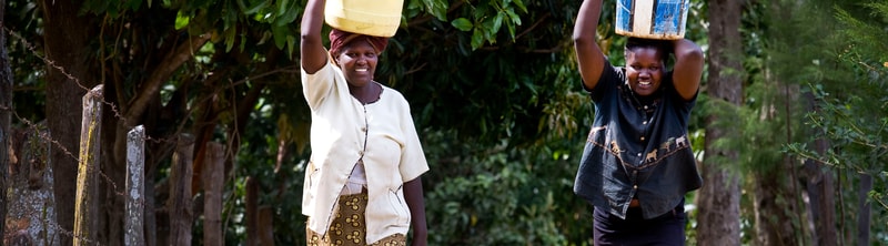 Two women walk along a rural path carrying large containers on their heads, smiling as they balance them above a fence and lush green trees.