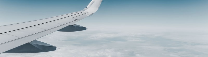 Wing of an airplane viewed from window, white metal surface with flaps and winglets, high above the clouds under a blue sky.