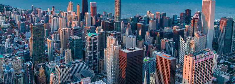 A dense city skyline with numerous tall skyscrapers, waterfront in the distance, illuminated by warm sunset light across a busy urban landscape.