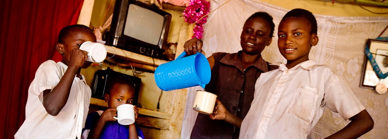 A group of four children share drinks in a cozy room; one child pours from a blue pitcher into a metal mug while another sips from a white cup, with a small child nearby and two others smiling.