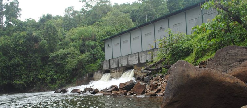 A long, light gray building with small doors sits on a rocky riverbank, a small waterfall spilling over concrete below, surrounded by lush green trees.