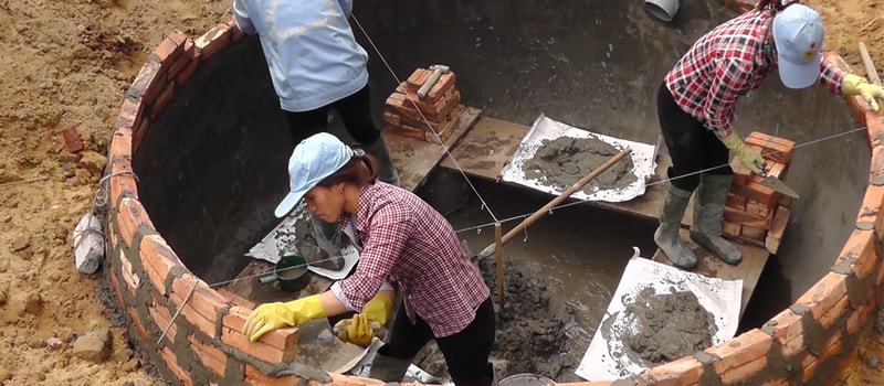 People building a circular brick structure in a muddy excavation site, laying bricks, wearing gloves and hats, with trowels and buckets inside the partial clay wall.