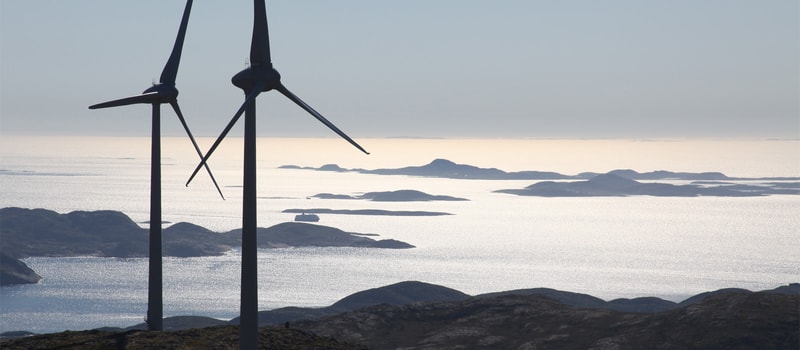 Two large wind turbines stand on a rocky coastal ridge, their blades partially visible, overlooking a sparkling calm sea with small islands dotting the horizon under a pale sky.