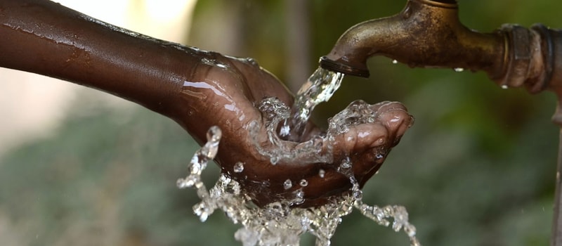 Hands under a running faucet, water splashes and streams around wrists, catching light as clear droplets cling to dark skin; rusty taps and green background blur.