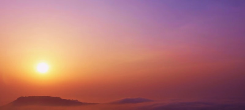 silky sunrise over a hazy horizon, warm orange and pink tones gradually fading to purple, distant silhouette of a hill barely visible through low clouds