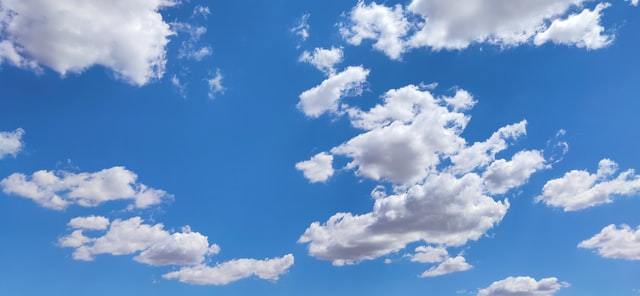 Blue sky with scattered white, fluffy cumulus clouds drifting across the horizon, bright and clear with ample sunlight.
