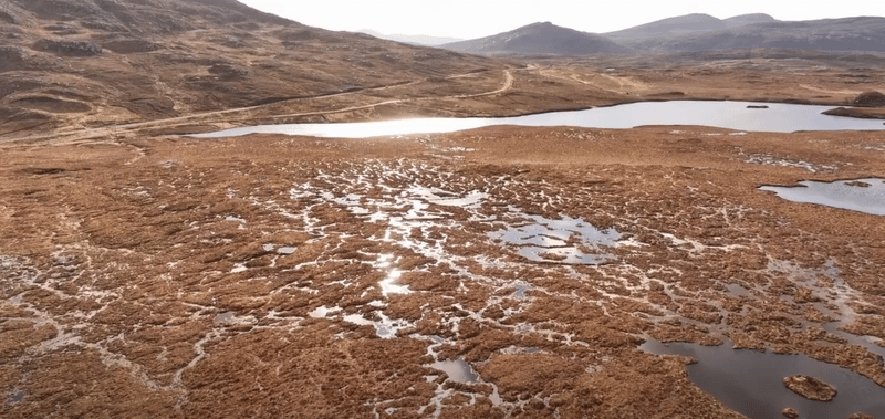 Arid, rolling hills with a shallow, reflective lake and winding dirt path; brown, mossy terrain stretches toward distant mountains under a clear sky.