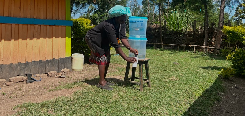 A person wearing a turquoise head covering fills a bottle at a water dispenser placed on a wooden stool, outdoors near a colorful wooden wall and green yard.
