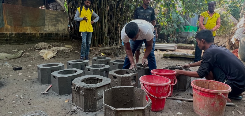 Workers build concrete blocks outdoors, shaping cement around hollow cylindrical forms; several red buckets sit nearby, while onlookers in yellow vests observe.