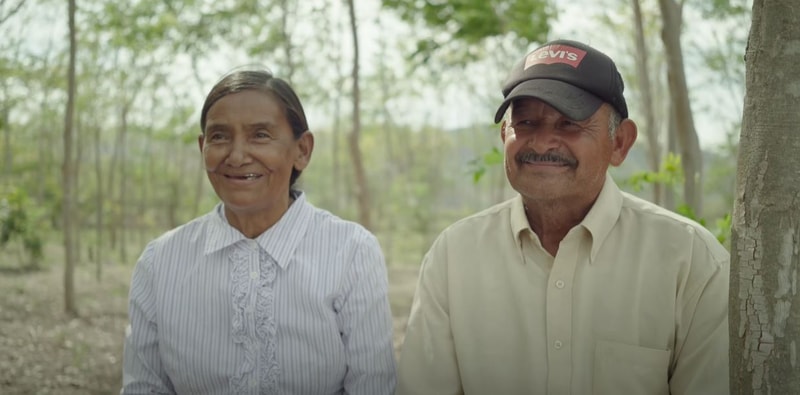 Two seniors sit outdoors among slim trees, smiling warmly; the woman wears a light striped blouse with lace front, and the man wears a beige button-up shirt and a black Levi's cap.